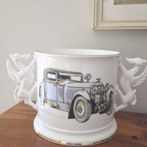 Peut inclure: Un récipient en céramique blanche doté de deux anses sculptées en forme de dragon et d'un bord et d'une base dorés. L'avant présente une illustration détaillée d'une voiture ancienne bleu clair et or avec le texte Bentley Speed Six 1930.