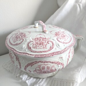 Puede incluir: Orinal de cer&aacute;mica blanca con tapa, con un estampado floral rosa. El orinal tiene un asa y un borde decorativo. La tapa tambi&eacute;n tiene un asa. El orinal est&aacute; sobre un tapete de encaje blanco.