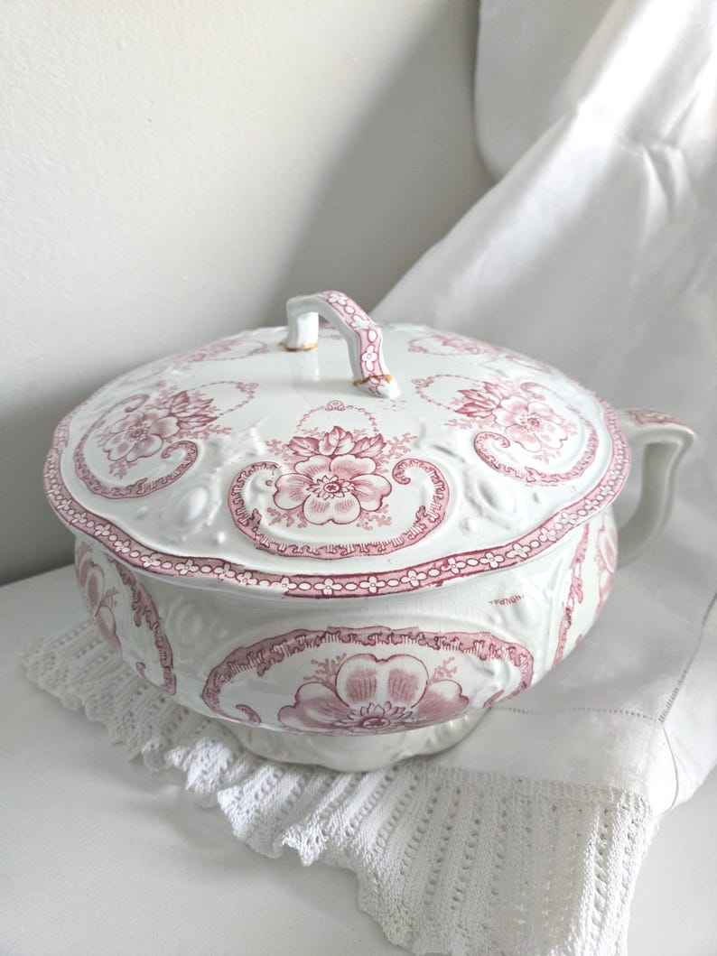 Puede incluir: Orinal de cer&aacute;mica blanca con tapa, con un estampado floral rosa. El orinal tiene un asa y un borde decorativo. La tapa tambi&eacute;n tiene un asa. El orinal est&aacute; sobre un tapete de encaje blanco.