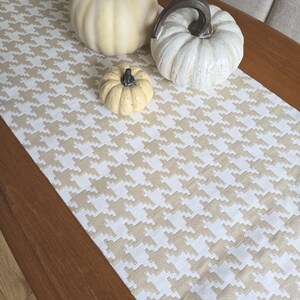 May include: A wooden table with a beige and white houndstooth patterned table runner. Three decorative pumpkins in cream and yellow hues are arranged on the runner. The pumpkins vary in size and shape, creating a fall-themed display.