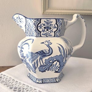 Może przedstawiać: Biały ceramiczny dzbanek o sześciokątnym korpusie i zakrzywionym uchwycie. Powierzchnia ozdobiona jest misternymi niebieskimi wzorami, w tym centralnym motywem pawia, elementami kwiatowymi oraz geometrycznymi obwódkami przy krawędzi i podstawie.