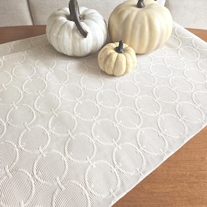 May include: Three decorative pumpkins of different sizes rest on a cream-coloured table runner with a repeating circle pattern. The pumpkins are white and cream, with brown stems. The runner is on a wooden surface.