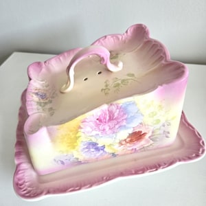 May include: A vintage pink ceramic butter dish with a floral design. The rectangular dish has a lid with a handle and is decorated with pink, blue, and yellow flowers. The dish sits on a matching tray with a scalloped edge.