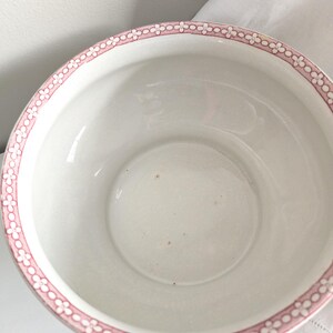 Puede incluir: Un cuenco de cer&aacute;mica blanca con un borde floral rosa. El cuenco tiene un dise&ntilde;o amplio y abierto y un interior liso. El borde rosa presenta un patr&oacute;n repetido de peque&ntilde;as flores estilizadas. El cuenco es probablemente vintage o antiguo.