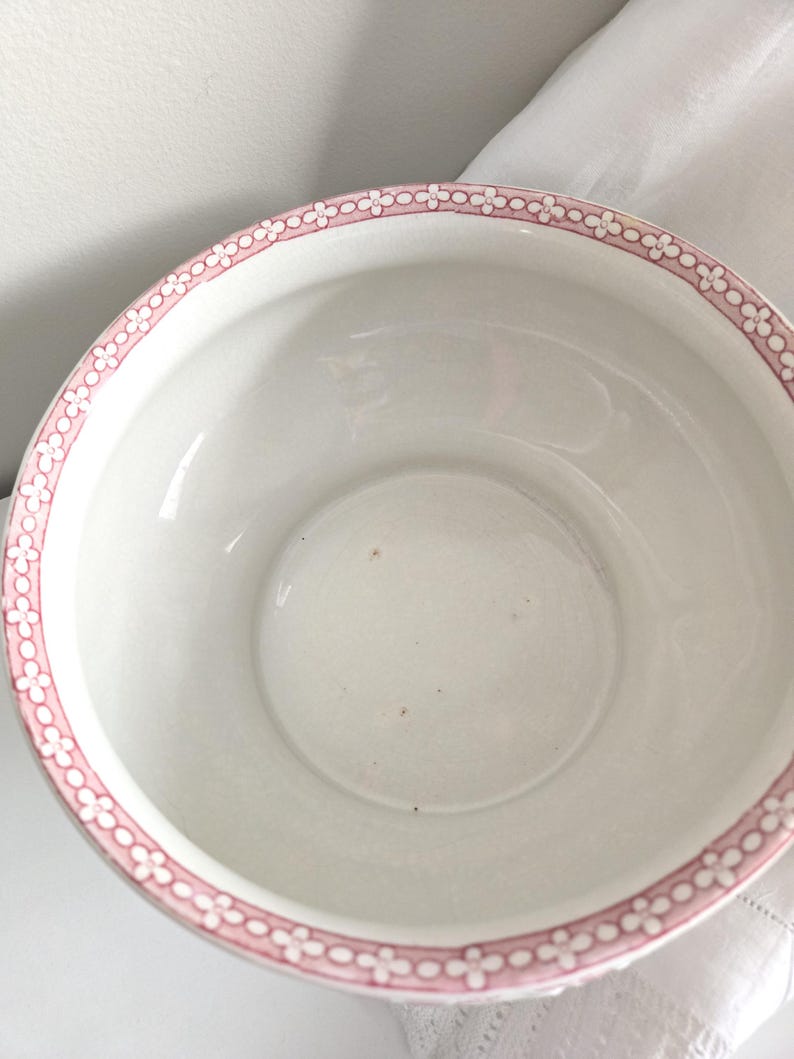 Puede incluir: Un cuenco de cer&aacute;mica blanca con un borde floral rosa. El cuenco tiene un dise&ntilde;o amplio y abierto y un interior liso. El borde rosa presenta un patr&oacute;n repetido de peque&ntilde;as flores estilizadas. El cuenco es probablemente vintage o antiguo.