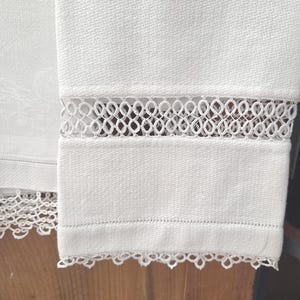 Puede incluir: Una toalla de lino color blanco roto con un tejido texturizado, una banda horizontal de intrincado encaje calado y un delicado borde de encaje festoneado a lo largo del dobladillo inferior, mostrando un diseño textil limpio.