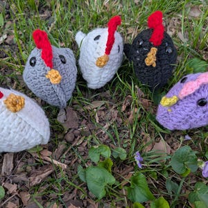 May include: Five handmade crochet chicken toys in various colors: white, gray, black, and purple. Each chicken has a red comb, black eyes, and a yellow wattle. The toys are set on a bed of grass and leaves.