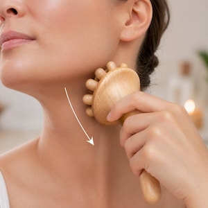 Puede incluir: Una herramienta de masaje de madera con una cabeza redondeada y múltiples perillas salientes se utiliza en el cuello. La herramienta se sostiene en la mano y se mueve a lo largo del cuello, siguiendo una flecha hacia abajo. La herramienta es de madera clara.