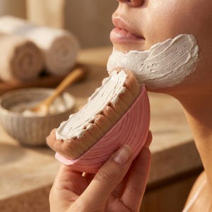 Può includere: Una spazzola per il viso rotonda di colore rosa con setole morbide marrone chiaro ricoperte da una spessa maschera all'argilla bianca. La spazzola è tenuta contro il mento, applicando la crema bianca in uno strato liscio. Il manico è rosa e rigato.
