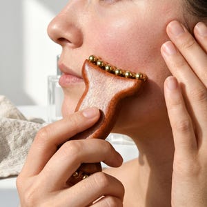 Pode incluir: Uma ferramenta de massagem facial de madeira marrom com bolas de metal douradas está sendo usada na bochecha de uma pessoa. A ferramenta tem um formato único. A mão da pessoa segura a ferramenta, e a outra mão toca seu rosto.