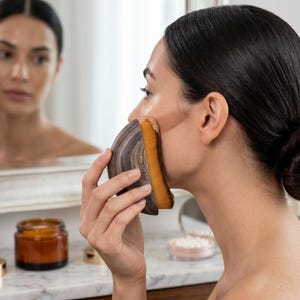 Pode incluir: Uma mulher aplicando maquiagem com um pincel com cabo de madeira. O pincel tem um padrão de grão de madeira escura e uma cabeça de pincel de cor bronzeada. Um pote de creme e um estojo compacto são visíveis ao fundo.