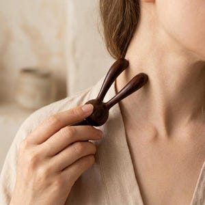 Könnte beinhalten: Ein dunkelbraunes Massagewerkzeug aus Holz mit einem abgerundeten Mittelteil und zwei länglichen, geschwungenen Zinken, die nach außen ragen. Die glatte, polierte Oberfläche des Holzes betont die natürliche Maserung und die tiefe Farbe.