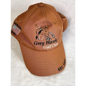 May include: A brown baseball cap with the text "Grey Hawk Golf Club" and a graphic of a hawk. The cap also features the American flag and the text "AGRAN, OHIO" on the side, with "EST. 2004" on the brim.