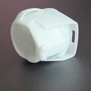 May include: A light blue, geometric-shaped object with a rounded base and a rectangular top. The object has a small, rectangular opening on one side and two small, circular protrusions on the front. The object appears to be made of plastic.