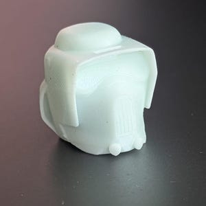 May include: A light green, 3D-printed object resembling a helmet or headgear. The object has a rounded top, a visor-like brim, and a detailed front section. The overall design suggests a futuristic or sci-fi theme.