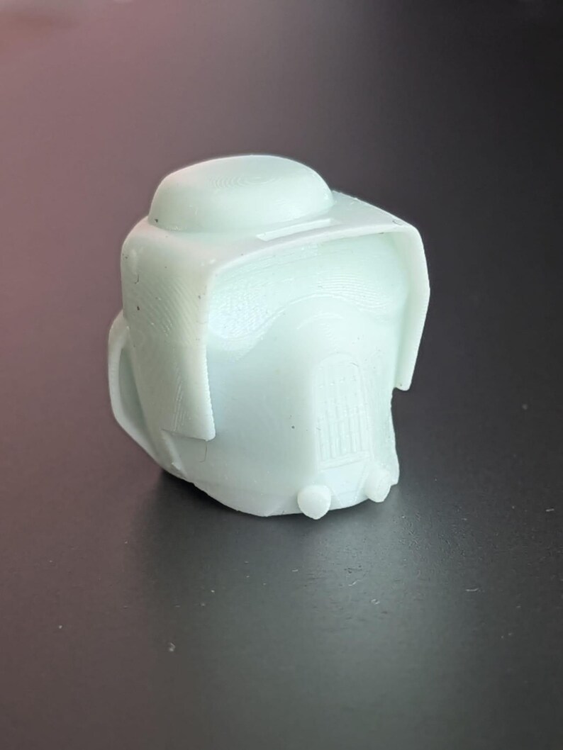 May include: A light green, 3D-printed object resembling a helmet or headgear. The object has a rounded top, a visor-like brim, and a detailed front section. The overall design suggests a futuristic or sci-fi theme.