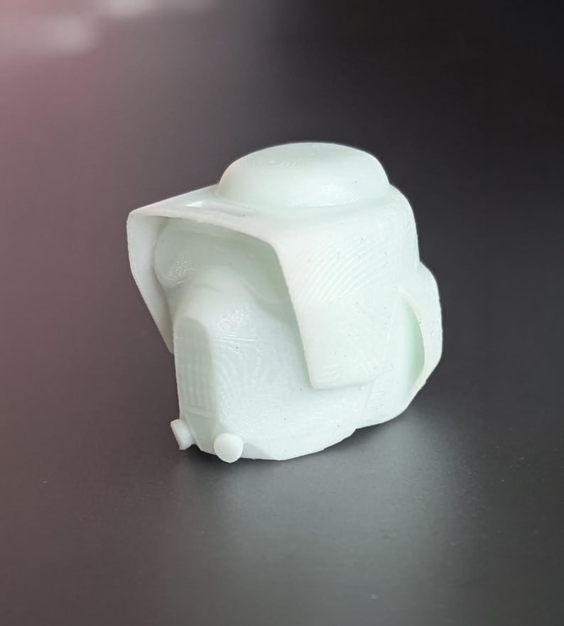 May include: A light green, 3D-printed helmet with a rounded top and a visor-like brim. The helmet has a textured surface and a detailed front, suggesting a futuristic or sci-fi theme. The object is set against a dark background.