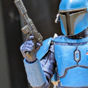 May include: A close-up of a blue Mandalorian action figure. The figure is wearing blue armour and a helmet with a black visor. The figure is holding a black blaster gun.