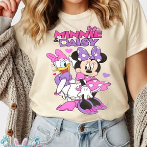 May include: Cream-colored t-shirt featuring Minnie Mouse and Daisy Duck. The graphic includes the text "Minnie & Daisy" in pink and purple, with the characters in their signature outfits. Pink hearts are scattered around the characters.