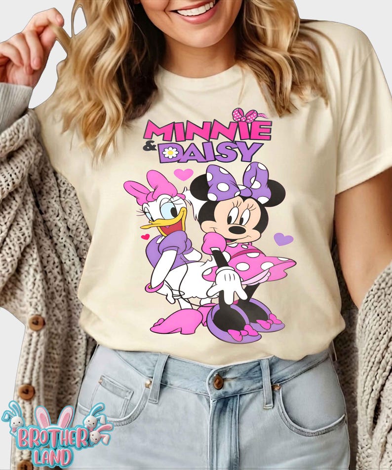 May include: Cream-colored t-shirt featuring Minnie Mouse and Daisy Duck. The graphic includes the text "Minnie & Daisy" in pink and purple, with the characters in their signature outfits. Pink hearts are scattered around the characters.