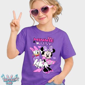 May include: Purple t-shirt featuring Minnie Mouse and Daisy Duck with the text "MINNIE & DAISY". The cartoon characters are in pink, white, and black. The shirt is worn by a person wearing pink heart-shaped sunglasses.