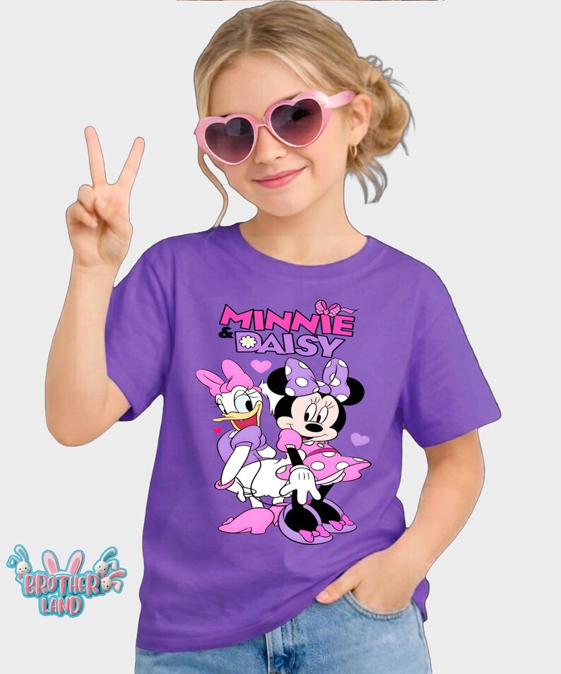 May include: Purple t-shirt featuring Minnie Mouse and Daisy Duck with the text "MINNIE & DAISY". The cartoon characters are in pink, white, and black. The shirt is worn by a person wearing pink heart-shaped sunglasses.