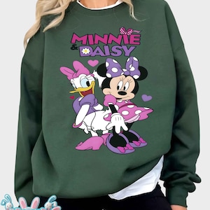 May include: Dark green sweatshirt featuring Minnie Mouse and Daisy Duck. The graphic includes the text "MINNIE & DAISY" in pink and purple, with a daisy and hearts. The characters are in their signature outfits.