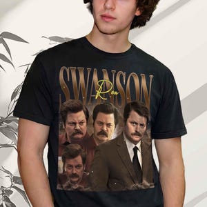 May include: A black short-sleeved t-shirt featuring a graphic print of three images of a man with a moustache. The text "SWANSON" is printed in large gold letters at the top, with the word "Ron" written in a cursive yellow script below it.