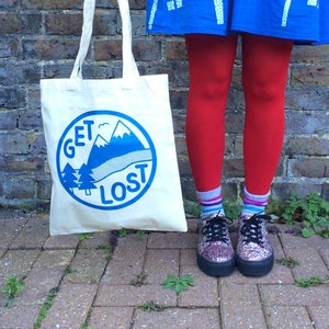 May include: A white canvas tote bag with blue text that reads "GET LOST" and a blue mountain scene inside a circle. The bag is being held by a person wearing a yellow shirt, a blue skirt, and red tights.