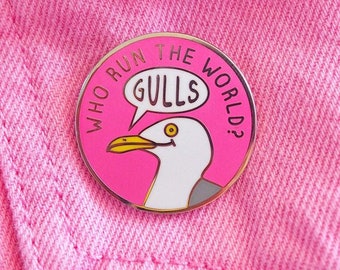 May include: Pink enamel pin with a white seagull and the text "Who run the world? Gulls." in a speech bubble.
