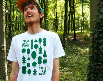 May include: A person wearing a white t-shirt with a green print of various trees and the text "Trees are good!" in a forest setting.