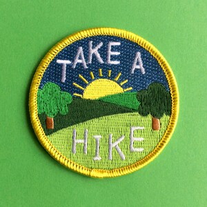 Peut inclure: Écusson brodé circulaire avec un paysage. Il présente un ciel bleu, un soleil jaune, des arbres verts et l'inscription "TAKE A HIKE" en blanc. Le patch a une bordure jaune et est posé sur un fond vert.