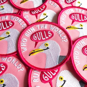 May include: A close-up of several pink embroidered patches featuring a white seagull and the text "Who Run The World? Gulls" in a speech bubble.