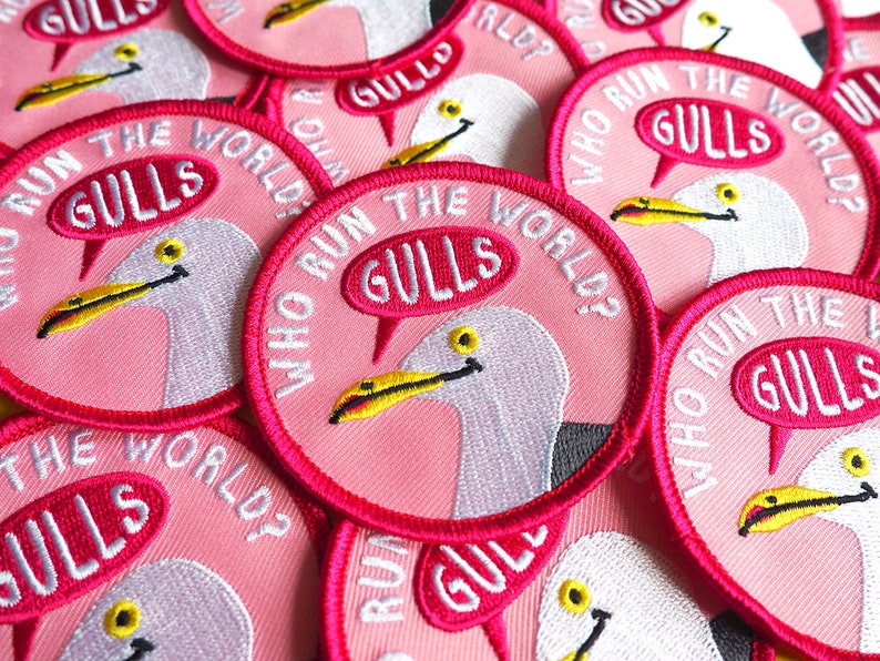 May include: A close-up of several pink embroidered patches with a white seagull and the text "Who Run The World? Gulls" in a speech bubble.