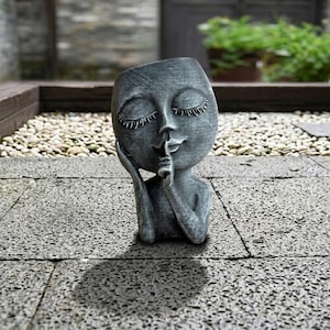 May include: A gray, stylized sculpture of a person with closed eyes, finger to lips, in a 'shush' gesture. The sculpture has a smooth face and is set on a stone surface. The sculpture is a decorative item.
