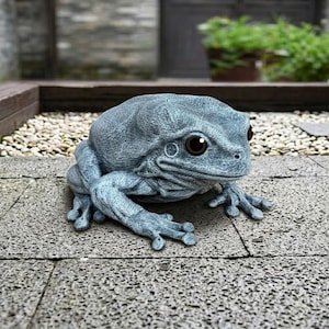 Peut inclure: Une statue de grenouille détaillée, bleu-gris, avec de grands yeux bruns. La grenouille est posée sur une surface en pierre, mettant en valeur sa peau texturée et ses détails. Cette statue de jardin décorative ajoute une touche fantaisiste aux espaces extérieurs.