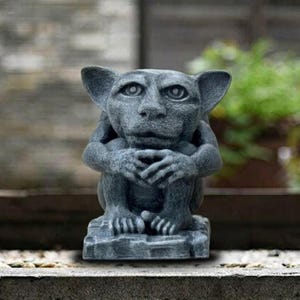 May include: A gray gargoyle statue with large ears, sitting with its hands clasped. The statue has a detailed face and is made of a stone-like material. The gargoyle is on a square base, and the background is blurred.