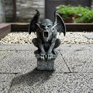 May include: A grey gargoyle statue with an open mouth and large wings. The statue is perched on a square pedestal. The gargoyle has a detailed, textured surface and is positioned outdoors on a stone patio.