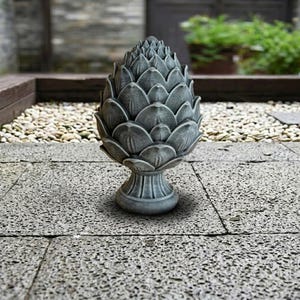 May include: A gray, stone-like artichoke sculpture with detailed petal-like layers. The sculpture sits on a pedestal base and is placed on a stone patio. The background features a blurred outdoor setting with greenery and small pebbles.