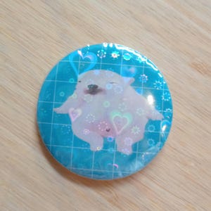 May include: A circular badge featuring a cartoon dog with closed eyes, set against a blue background with hearts and floral designs. The dog is white and fluffy. The badge has a holographic sheen.