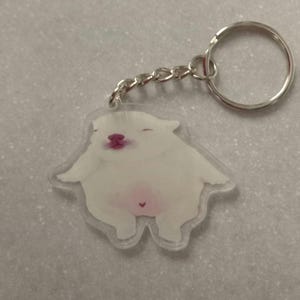 May include: A clear acrylic keyring featuring a printed illustration of a plump white animal with closed eyes, a pink nose, and a small pink heart marking on its belly. The charm is attached to a silver-toned metal split ring by a short chain.