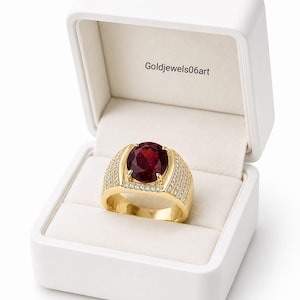 May include: A gold ring featuring a large, oval-cut deep red gemstone set in the center, flanked by rows of small, clear pavé-set stones along the band. The ring is displayed inside an open white jewelry box with the text Goldjewels06art printed inside.
