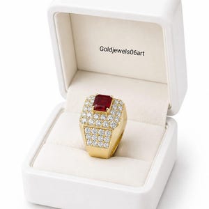 May include: A gold ring featuring a rectangular red gemstone at the center, surrounded by multiple small clear stones on the band and setting. The ring is displayed inside a white jewelry box with the text Goldjewels06art printed on the interior lid.