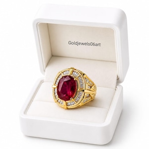 May include: A gold ring featuring a large oval-cut red gemstone surrounded by a halo of baguette-cut diamonds, set in a yellow gold band with intricate geometric detailing, presented inside a white jewelry box with the text Goldjewels06art.