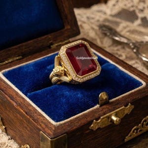 May include: A gold ring featuring a large, rectangular, deep red gemstone set in a halo of small white stones. The band displays intricate lion head detailing on the sides. The text Goldjewels06art is visible on the surface of the red stone.