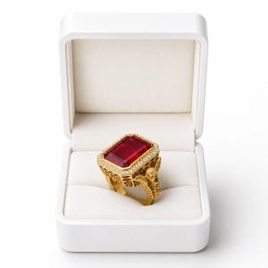 May include: A gold ring featuring a large rectangular red gemstone set in a halo of small white stones. The band and setting display intricate filigree metalwork with sculpted wing-like details on the sides, presented in a white jewelry box.