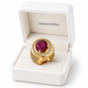 May include: A gold ring featuring a large, round, deep red gemstone at the center, surrounded by a halo of small clear stones. The band has a textured, open-work lattice design. The ring is displayed inside a white box with Goldjewels06art text.