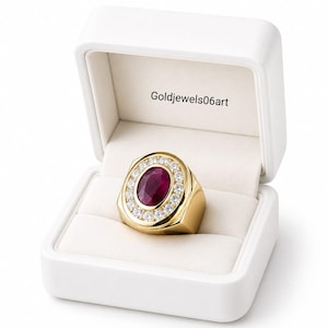 May include: A gold ring featuring a central oval-cut deep red ruby surrounded by a halo of small round white diamonds, presented inside a white jewelry box with the text Goldjewels06art printed on the interior lid lining.