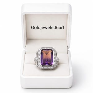 Op de afbeelding: Een zilveren ring met een grote, rechthoekige edelsteen met een kleurverloop van paars naar oranje, omringd door een halo van kleine witte steentjes. De ring wordt getoond in een open wit doosje met de tekst Goldjewels06art.