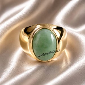 May include: A polished gold ring featuring a large, smooth, oval-shaped light green jade stone set in the center. The band is thick and rounded with a high-shine finish. The text Goldjewels06art is printed across the surface of the green stone.